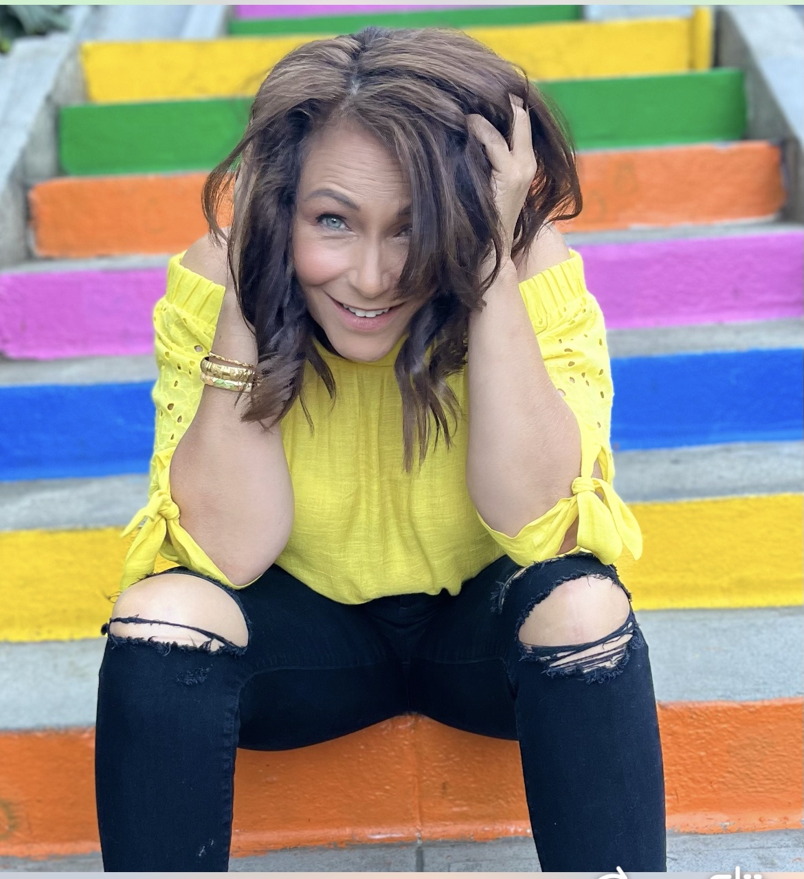Marie Finch smiling playfully in a yellow top and torn jeans on colorful stairs